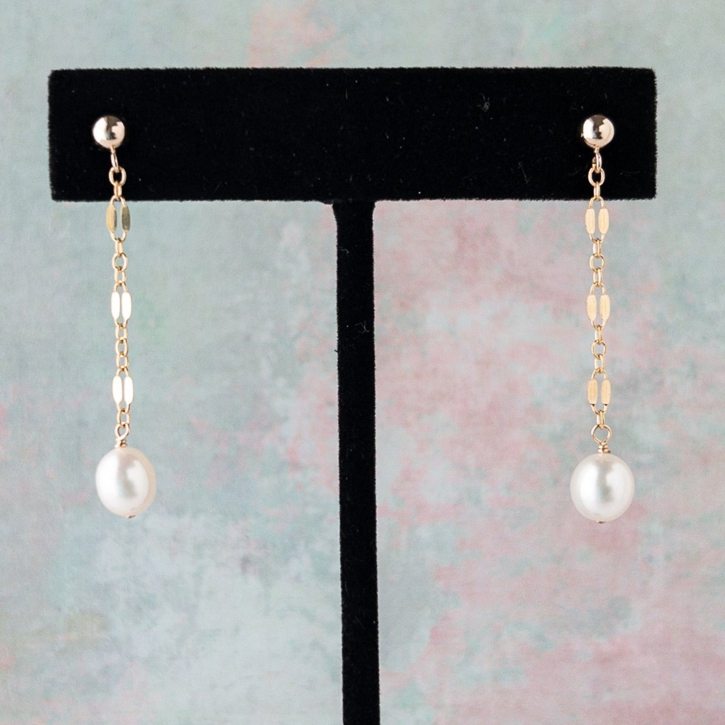Isabella Dangle Pearl earrings on a black earring stand against a pastel background