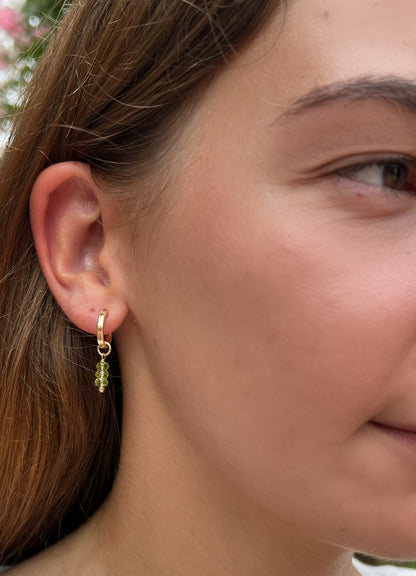 A close-up of a woman's ear wearing a small gold huggie hoop earring with a dangling green bead charm.