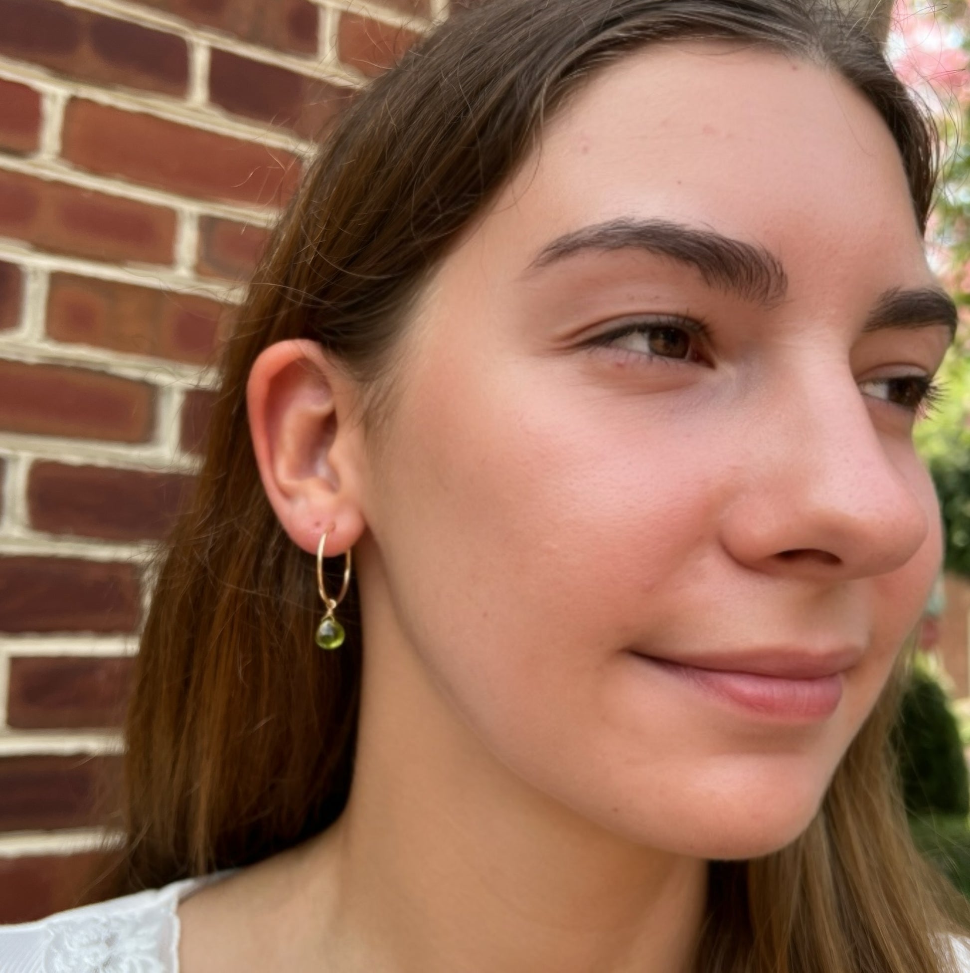 A close-up of a woman wearing a delicate gold hoop earring with a small green bead drop, set against a brick wall background.