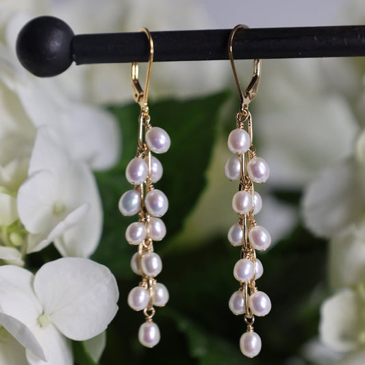 Gold drop earrings with clusters of white freshwater pearls hanging from a black display stand, with white flowers and green leaves blurred in the background.