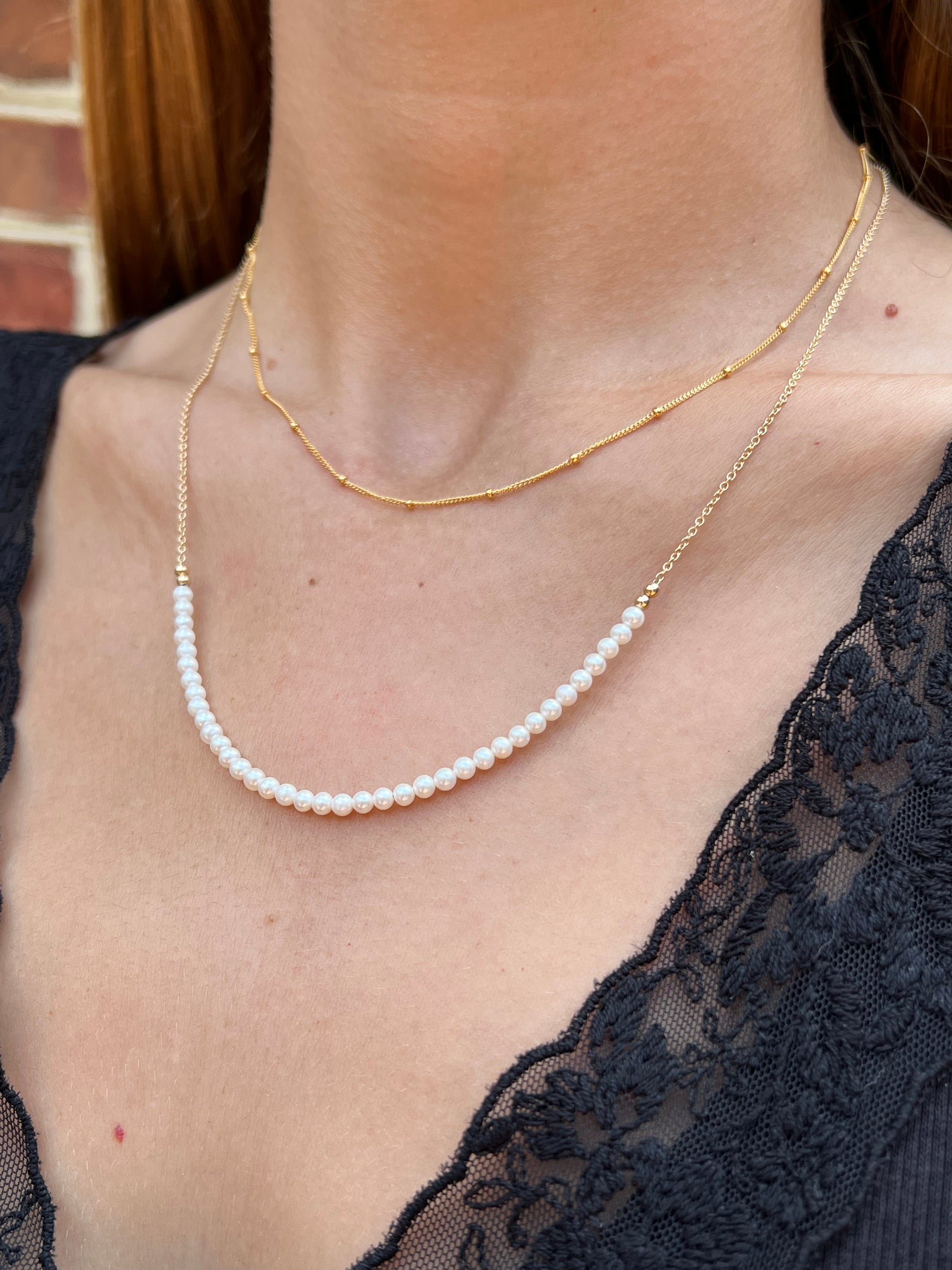 A close-up of a person's neck and upper chest wearing a delicate layered necklace. The necklace features a fine gold chain with a single strand of small white pearls ending in a small gold bead accent.
