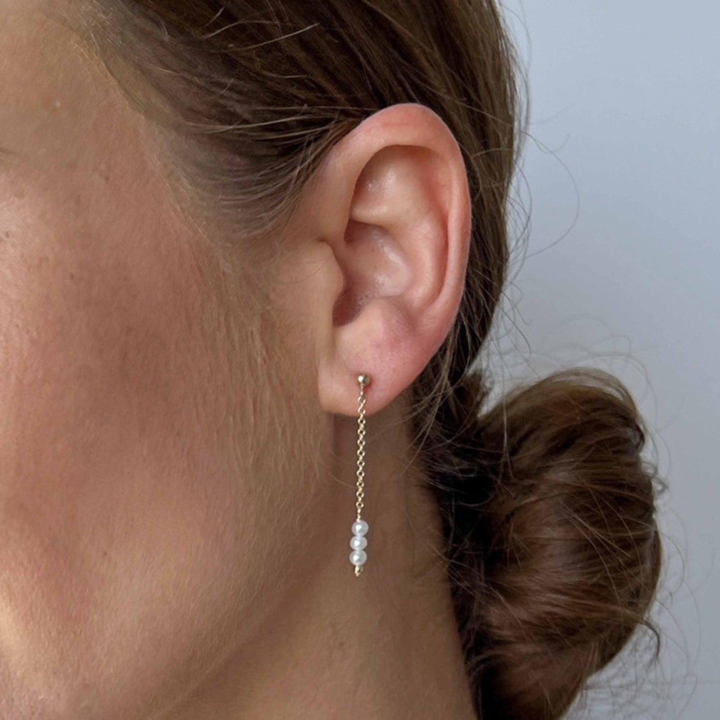 Close up of model wearing dainty pearl dangle earrings.  Tiny freshwater pearls hang from stud earrings and a chain for a total length of 1.5 inches.