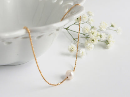 Gold necklace with a pearl pendant on a white surface with flowers and a cup.