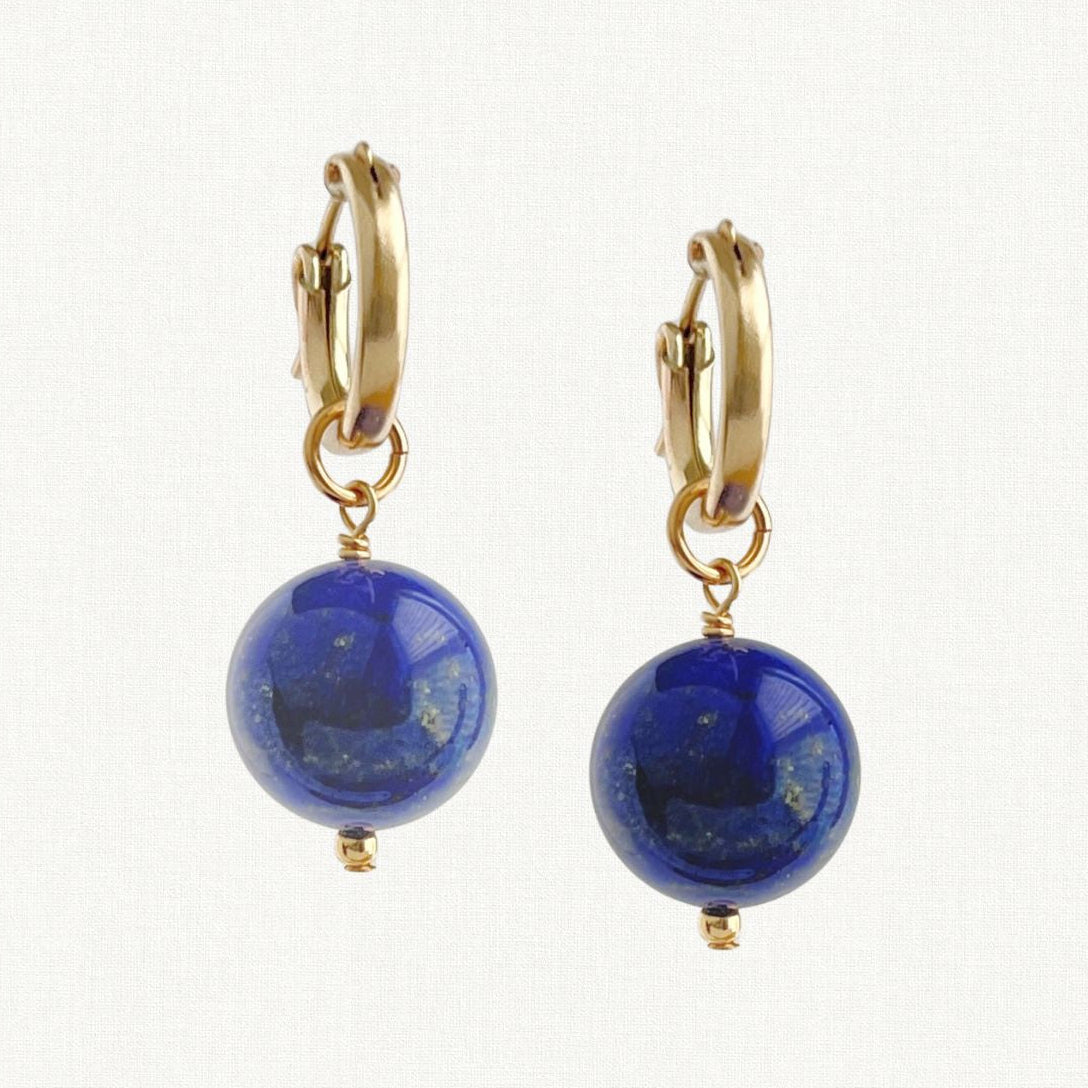 A pair of gold huggie hoop earrings with dangling polished lapis lazuli sphere beads, shown against a plain white background.