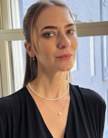 Woman wearing a black top and pearl necklace indoors with a window in the background